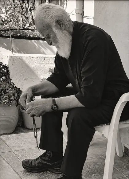Φωτογραφία: ηλικιωμένος άντρας με λευκή γενειάδα και κομπολόι, καθισμένος σε αυλή
