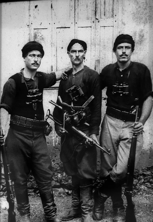 Φωτογραφία: τρεις άντρες με κρητικές φορεσιές και όπλα μπροστά σε ξύλινη πόρτα