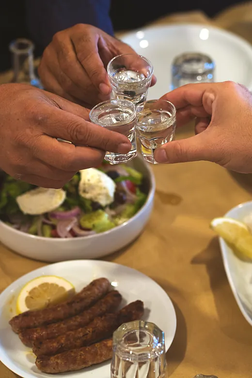 Δύο χέρια τσουγκρίζουν ποτηράκια ρακί πάνω από το στρωμένο τραπέζι